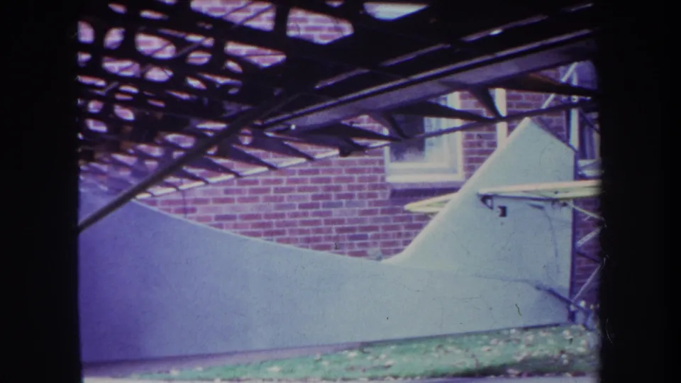 1969 8mm frame showing the white aircraft tail beneath a heavier pattern of overhead beams, with brick wall and grass visible.