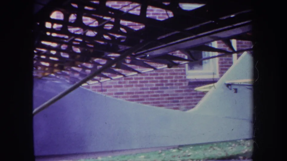 1969 Yucaipa home-movie frame showing a white custom aircraft body with exposed metal framework near a red brick building.