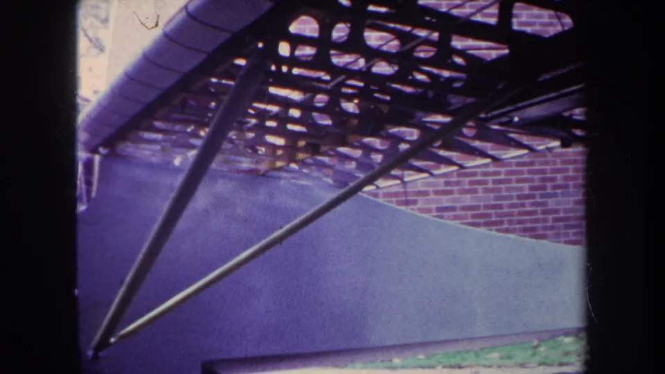 Handheld 8mm frame of the custom airplane under construction, emphasizing underside framework and diagonal supports against a brick wall.