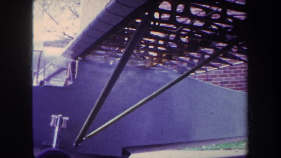 1969 home-movie frame in Yucaipa showing the aircraft side, diagonal brace, overhead framework, and a partial wheel or gear element.