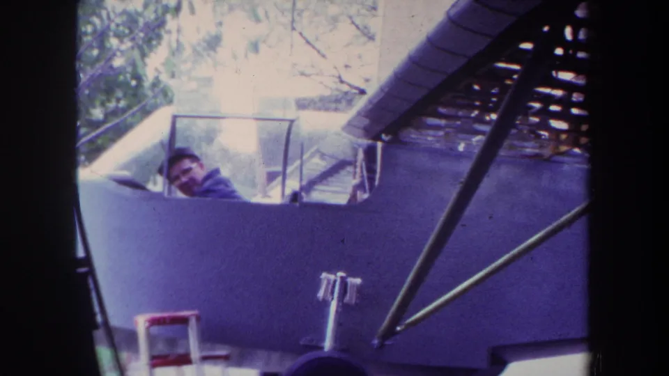 1969 Yucaipa home-movie frame showing an adult man by the cockpit of a purple custom airplane under construction.