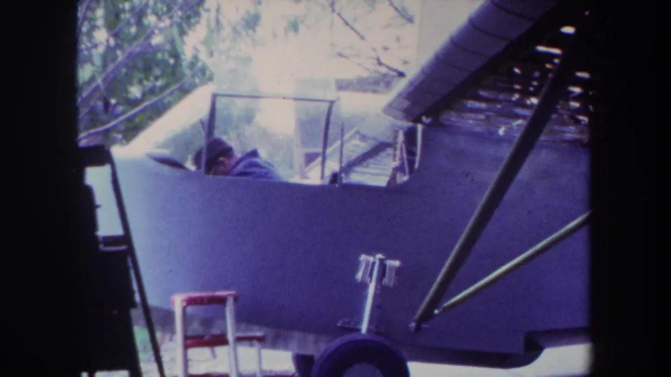 1969 Yucaipa frame of a pilot partly visible in a light single-engine airplane, with windshield, strut, and shaded structure behind.