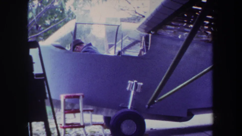 1969 Yucaipa home-movie frame showing the pilot lower in the cockpit of a light airplane, with exterior yard details around it.