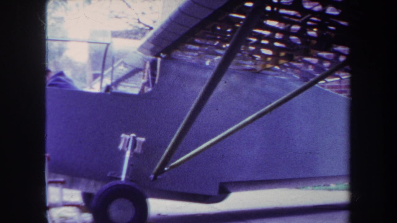 Close view of a small white airplane in 1969 Yucaipa footage, with fuselage, landing wheel, wing struts, and brick wall visible.