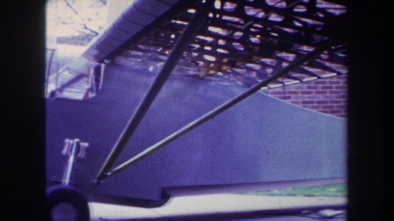 Parked small airplane with fuselage, struts, wheel, and brick wall in the background.