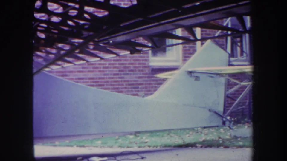 1969 Yucaipa home-movie frame showing a closer view of the small aircraft body against a brick residential wall.