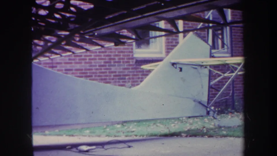 1969 Yucaipa frame showing the parked white aircraft beside a brick house, with fallen leaves and a cord visible near the pavement.