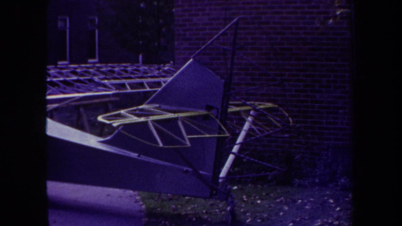 1969 home-movie frame from Yucaipa, California, showing a homemade aircraft with yellow-trimmed wing structure near a brick wall.