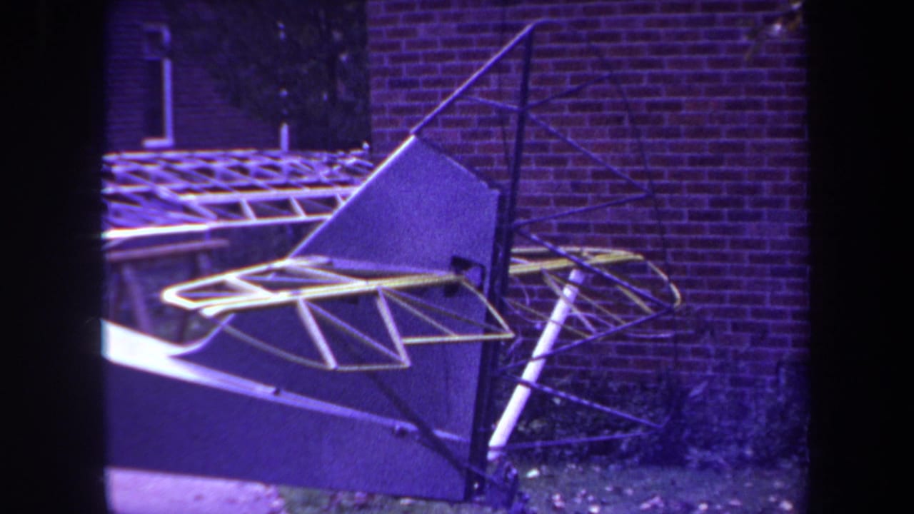 1969 Yucaipa 8mm frame showing a model airplane tail assembly with yellow metal framework beside a brick wall.