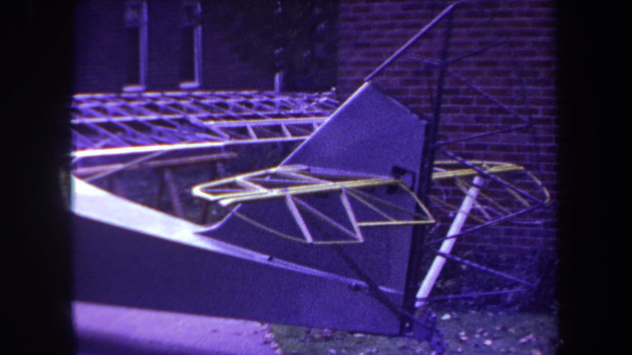 1969 Yucaipa home-movie frame showing a homemade sailplane or aircraft structure with yellow tubing beside a brick building.