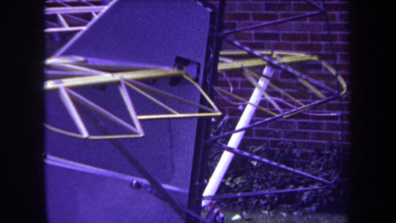 1969 Yucaipa home-movie frame showing a close view of a model aircraft tail assembly with metal rods, dark supports, brick, and foliage.