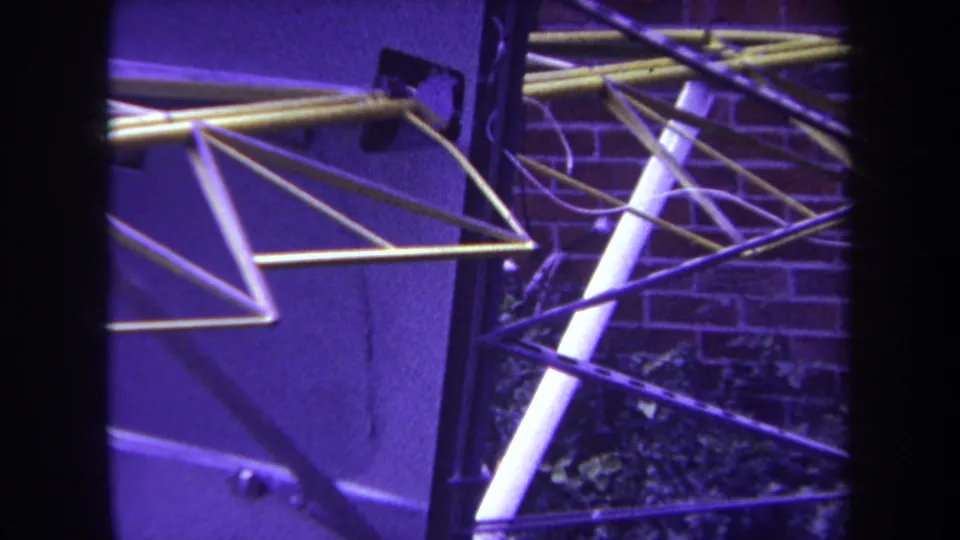 A 1969 Yucaipa 8mm frame shows the aircraft tail assembly from a nearby angle, with intersecting rods against brick and plants.
