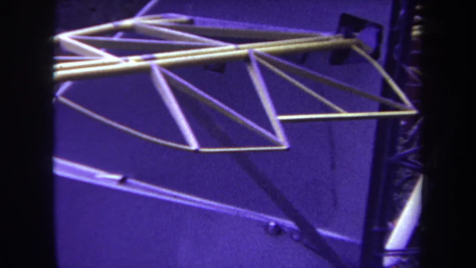 The camera moves across the aircraft tail frame, emphasizing triangular metal struts and rods over a purple-tinted surface.