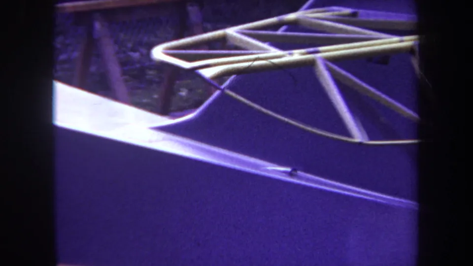 Final 1969 Yucaipa frame showing the aircraft tail structure from above a purple-tinted surface, with plants and dark supports behind it.