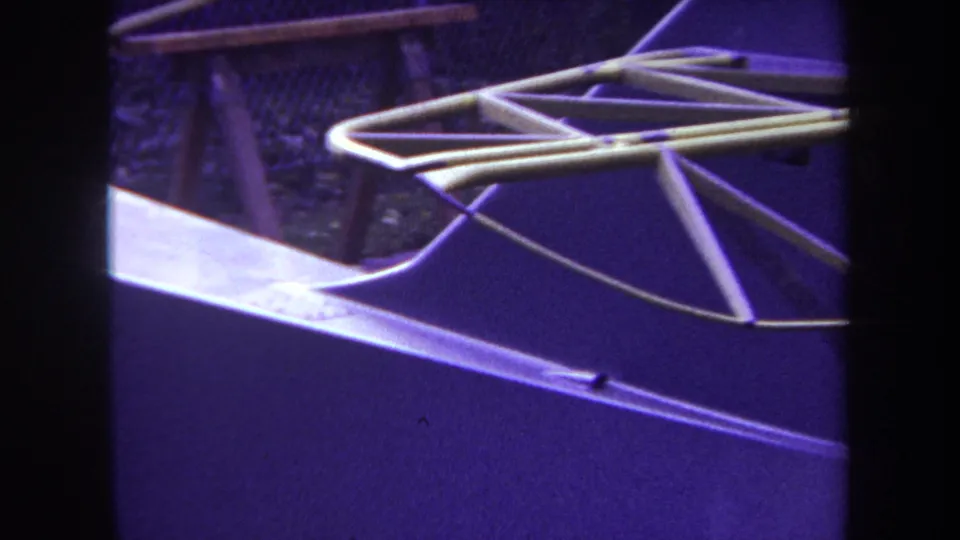 1969 Yucaipa-area 8mm frame with white metal rods forming geometric shapes over a purple-toned surface and chain-link fence.