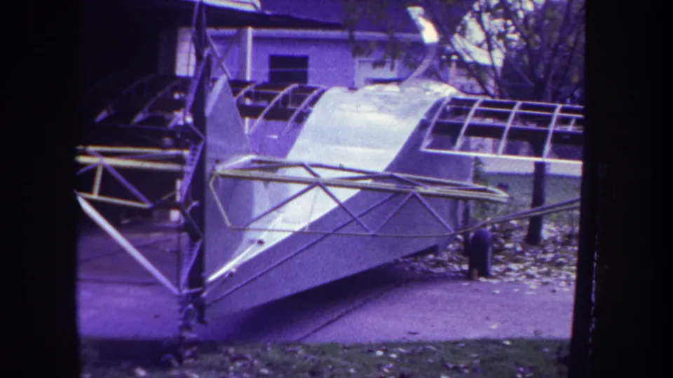 1969 Yucaipa home-movie frame showing the aircraft body and trusswork close to the camera, with driveway and foliage visible.