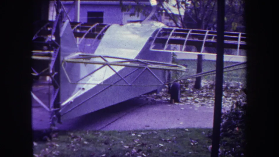 1969 home-movie frame of a hollow aircraft structure outdoors, with a small person visible near the fuselage.
