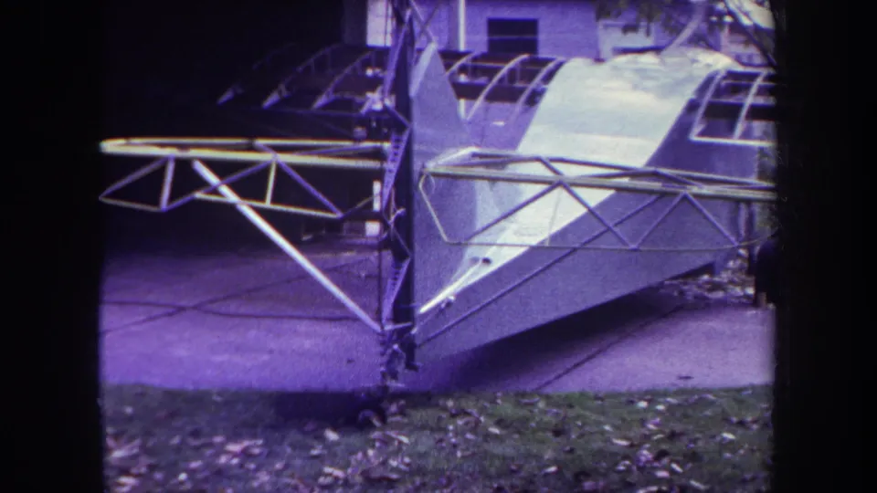 1969 Yucaipa home-movie frame showing an unfinished aircraft body with exposed tail framework, small landing wheel, driveway, grass, and a building behind it.