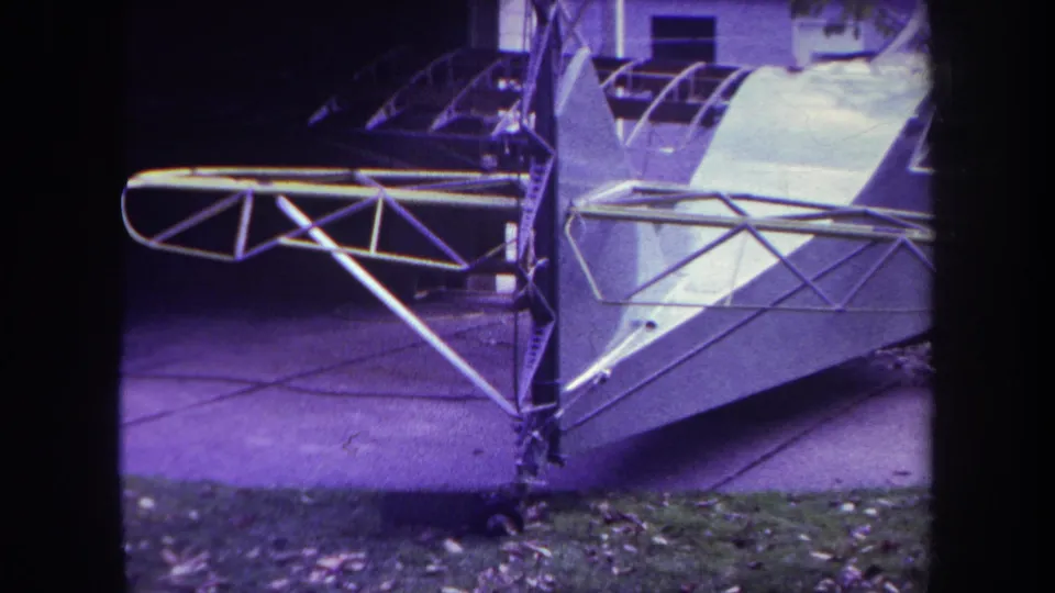 1969 Yucaipa footage frame showing the unfinished aircraft tail assembly, exposed tubing, fuselage skin, driveway, grass, and background building.