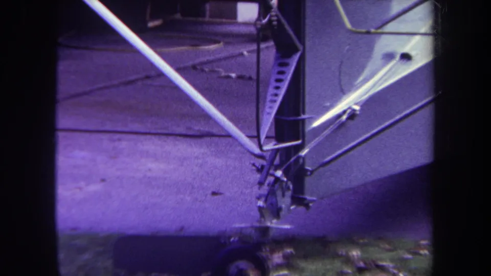 1969 Yucaipa home-movie frame showing a tight detail of the unfinished fuselage, metal support tubes, small wheel, pavement, and grass at the frame edge.