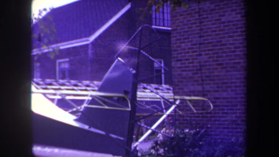 A purple-tinted 8mm view of an unfinished small aircraft resting beside brick residential buildings in the Yucaipa-area footage.
