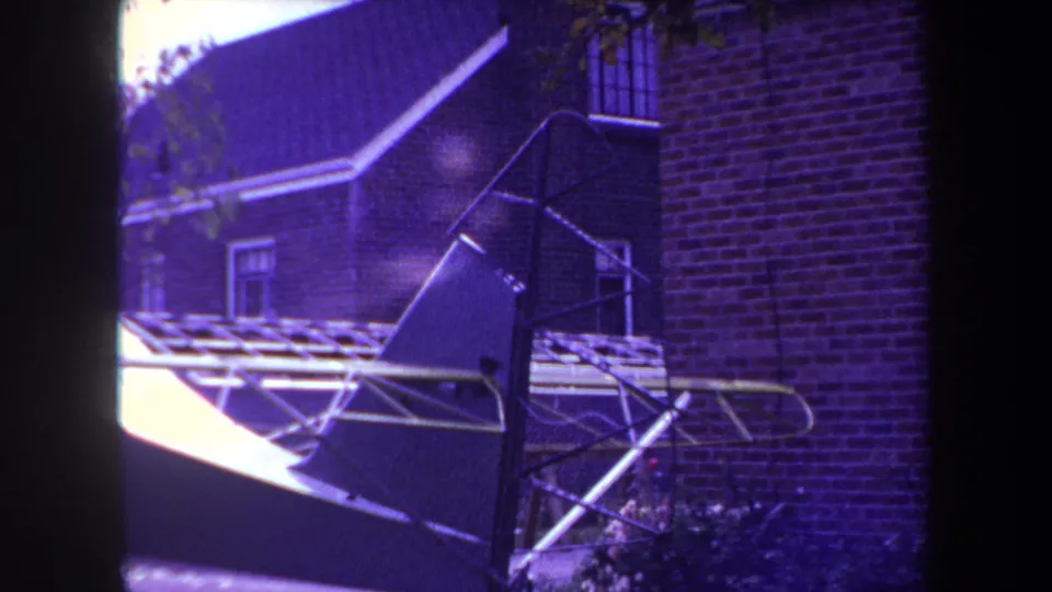 The frame shows the incomplete aircraft from another angle, with tubular supports, a brick wall, roof tiles, and a residential window visible.