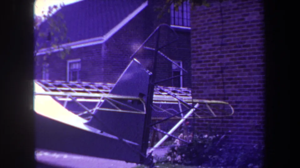A late-1960s 8mm frame shows the homemade aircraft structure stationary near brick walls, roof tiles, and low plants.
