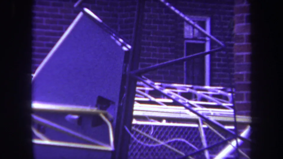 Purple-tinted 8mm frame from Yucaipa showing metal sculpture panels, rails, and a brick building background.