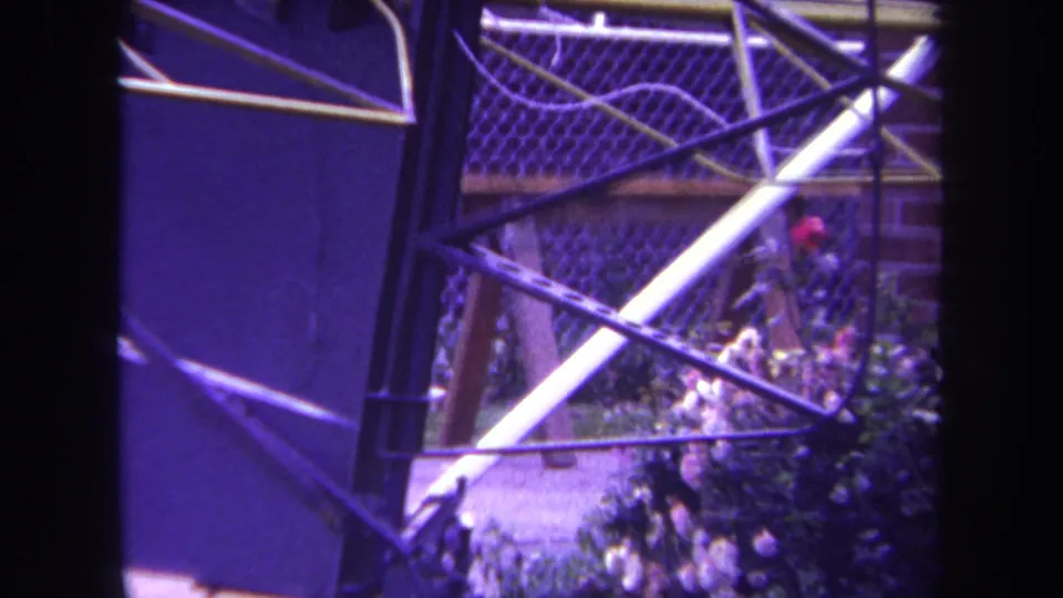 1969 Yucaipa home-movie frame showing a blue metal chain-link gate with diagonal bars, lawn edge, and garden planting.