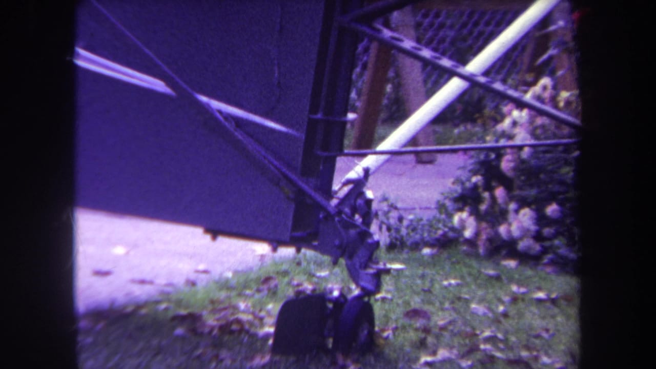 1969 Yucaipa frame with the lower corner of a blue chain-link gate, hinge area, grass, leaves, and sidewalk in view.