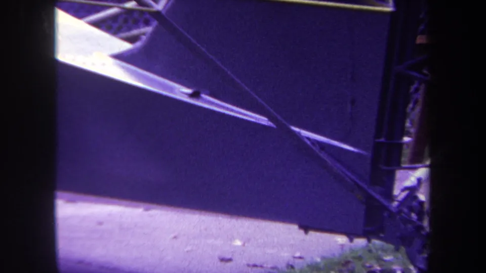 1969 Yucaipa home-movie frame showing an angled blue gate panel, metal braces, sidewalk, grass, and a few fallen leaves.