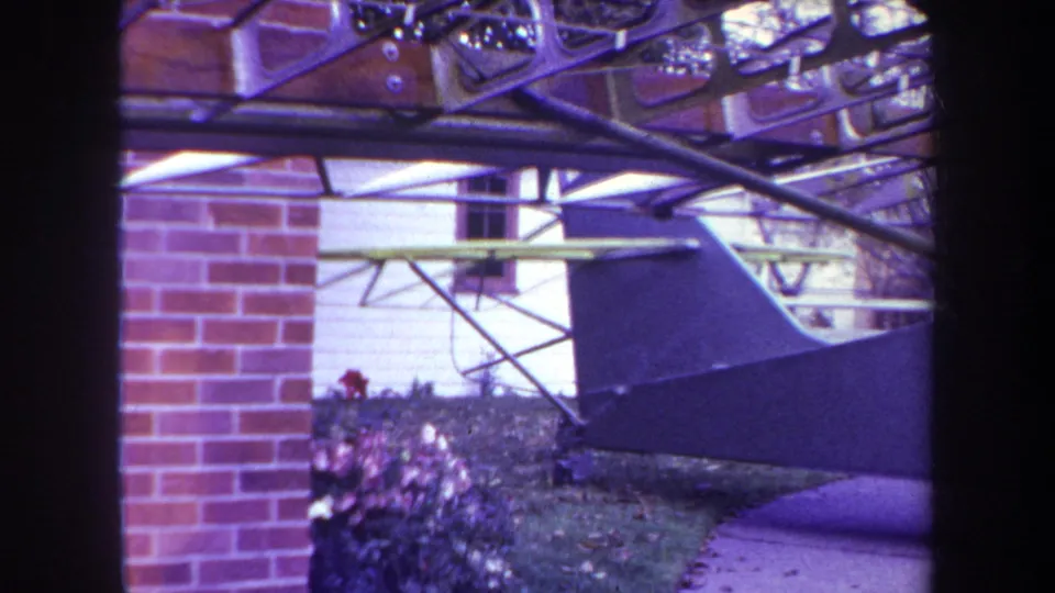 1969 Yucaipa home-movie frame showing an aircraft tail beside a white house wall, brick pillar, garden shrubs, and metal structure.
