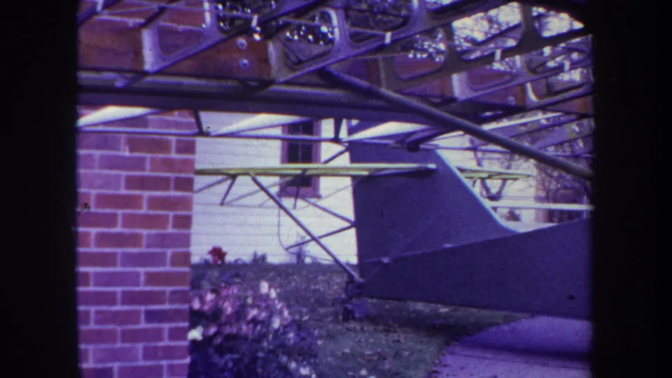 1969 Yucaipa frame showing the aircraft tail from a slightly shifted angle beside brick, siding, shrubs, and curved metal framing.