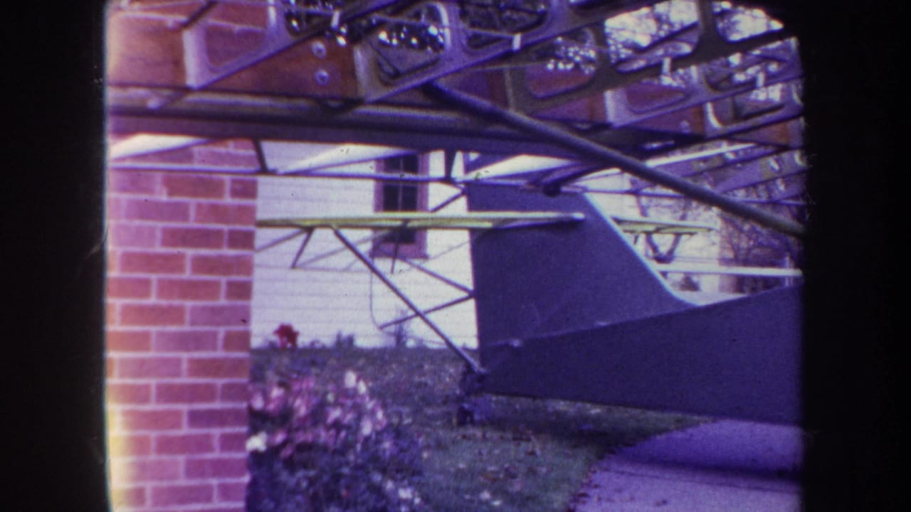 1969 8mm frame from Yucaipa showing part of a homemade airplane beside a brick wall, with exposed struts and a residential exterior behind it.