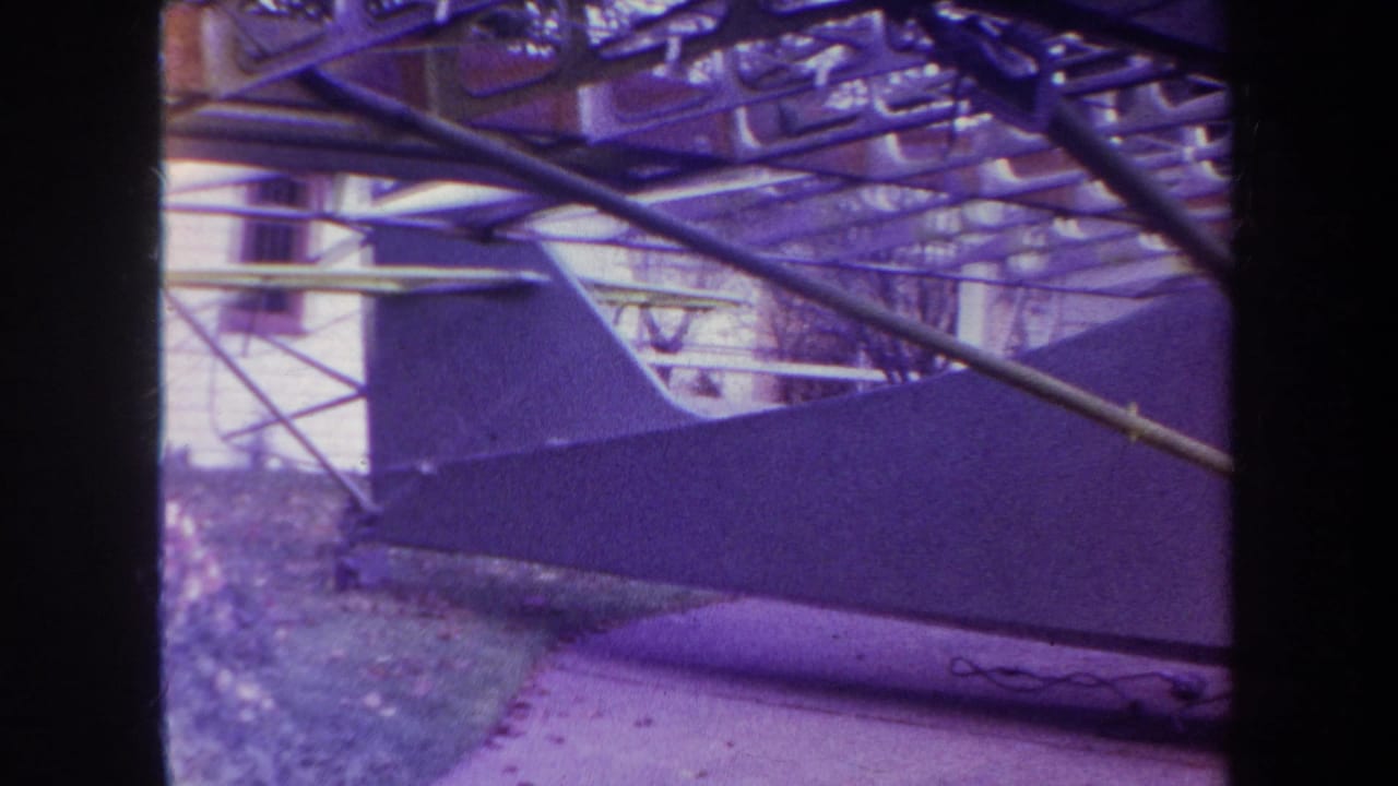 1969 8mm view of the airplane side and framework in a residential yard, with house siding and garden areas visible behind it.