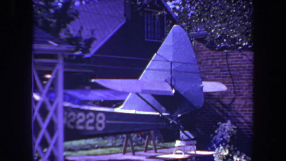 1969 8mm frame showing a homemade model airplane in a California backyard, with brick wall, lattice, and foliage behind it.