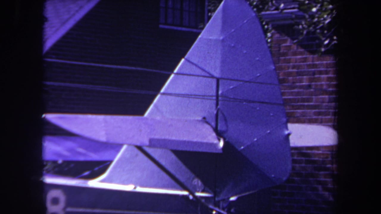 1969 8mm frame from Yucaipa, California showing a purple-tinted close view of a homemade airplane's triangular rudder and horizontal stabilizer near a brick residential wall.