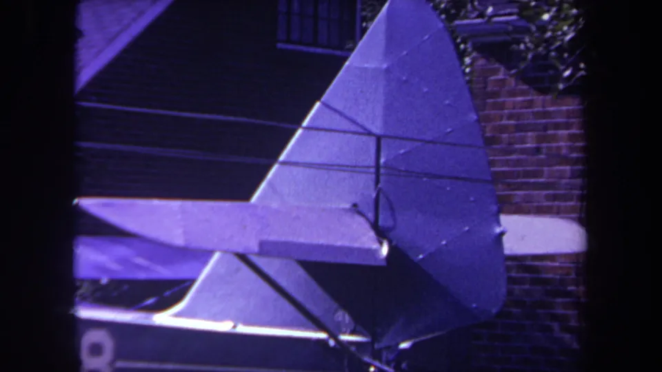 1969 8mm frame from Yucaipa, California showing a purple-tinted close view of a homemade airplane's triangular rudder and horizontal stabilizer near a brick residential wall.