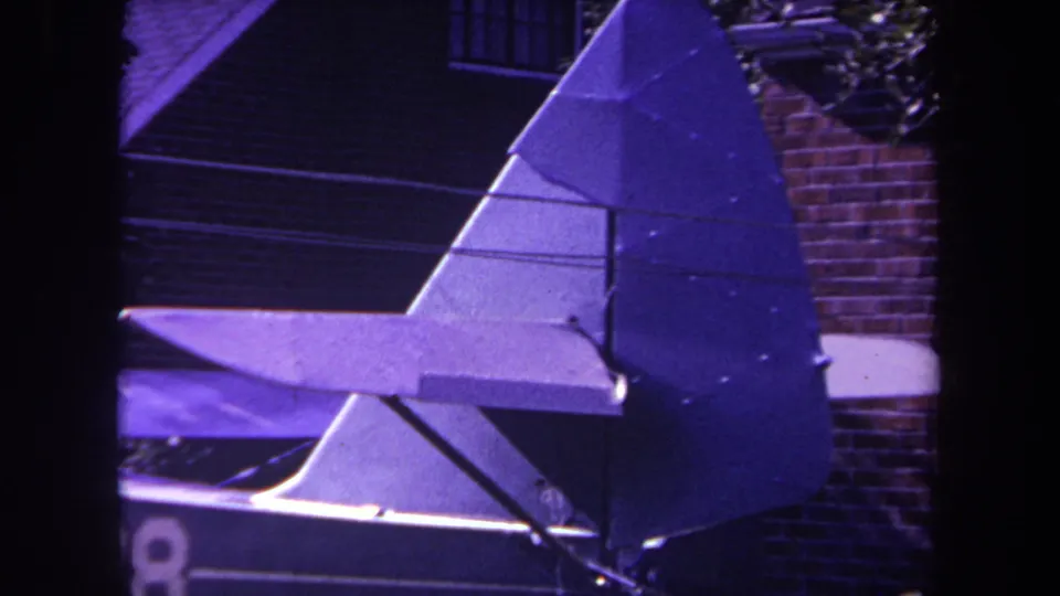 Purple-tinted 1969 home-movie frame showing the homemade aircraft tail assembly, including the rudder, stabilizer, and rear fuselage section, outside a house in Yucaipa, California.