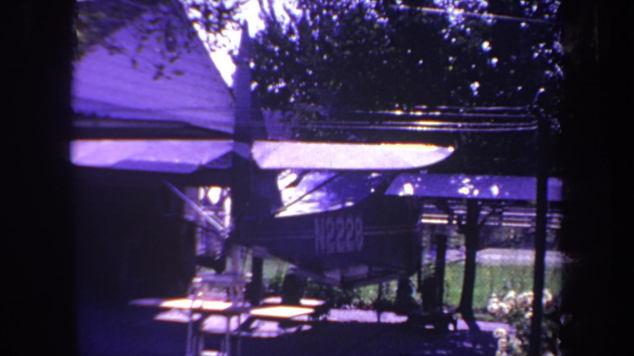 Small airplane parked under a shaded cover with trees and a house roof nearby.