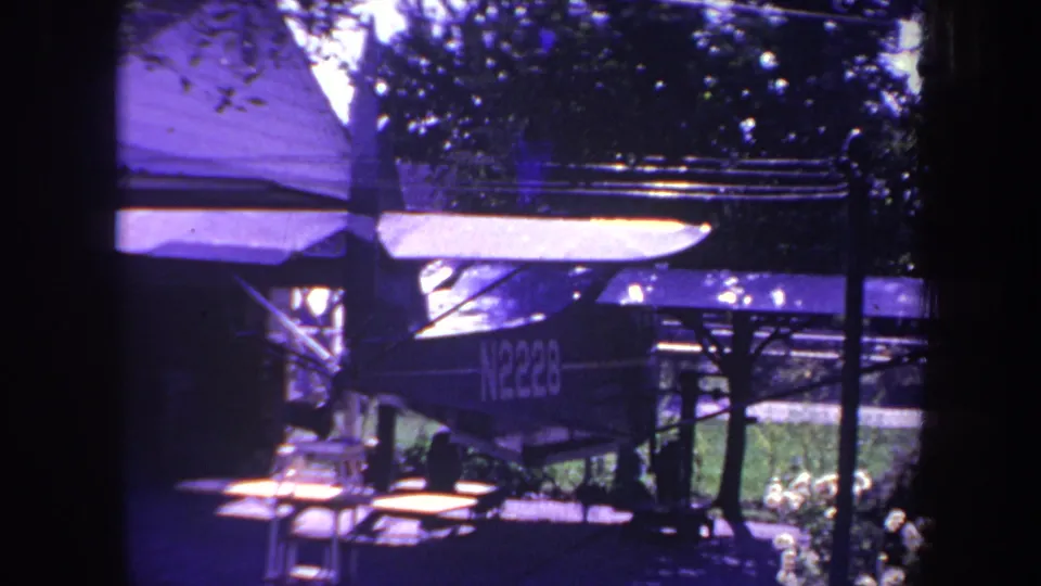 Aged 8mm frame from 1969 showing the parked airplane under cover, with foliage and structural posts around it.