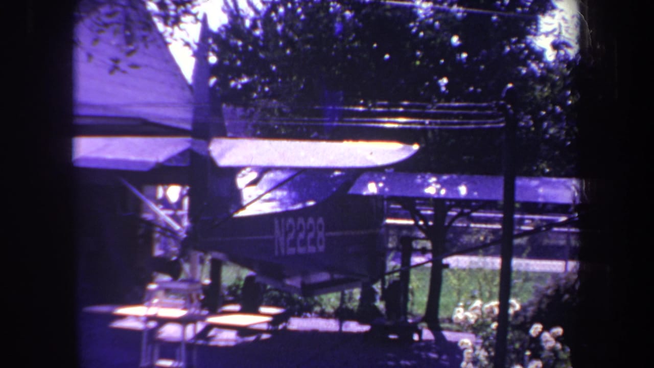 1969 Yucaipa home-movie frame showing a small single-engine aircraft parked beneath a shaded shelter in a residential yard, with a picnic table and trees nearby.
