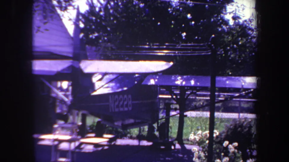 1969 Yucaipa frame with the same small aircraft seen through dark film-frame borders, parked under a simple shelter with greenery and a picnic table in the yard.