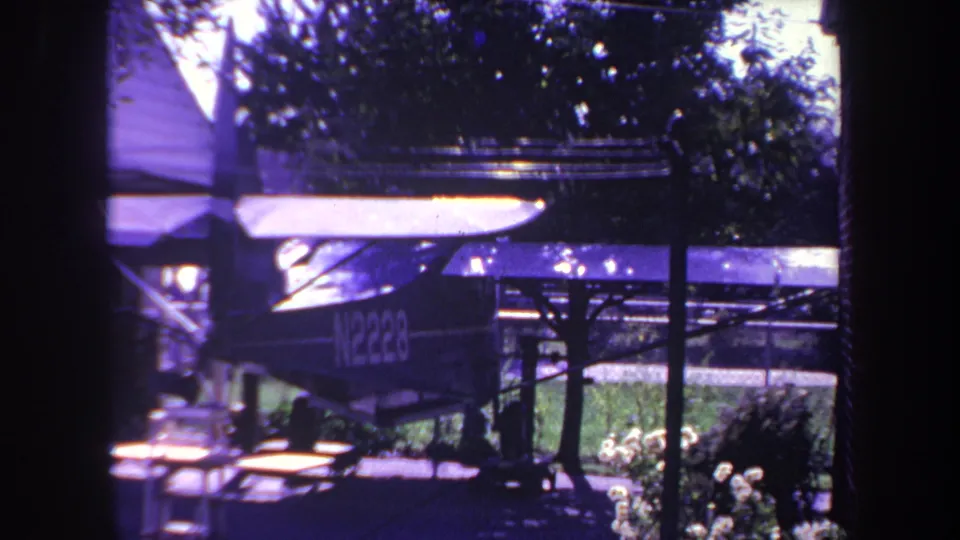 1969 Yucaipa home-movie frame showing the side of a small aircraft with visible registration markings, shaded by trees and positioned near a picnic table and garden plants.