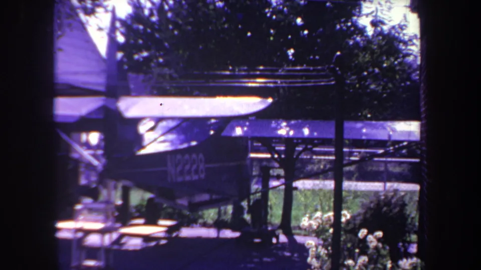 Final 1969 Yucaipa frame of the small aircraft beneath a makeshift yard shelter, with the picnic table, lawn, flowers, and dense tree shade visible around it.