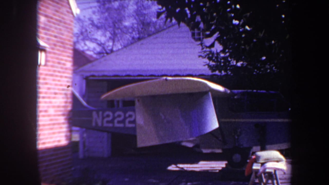1969 8mm home-movie frame from Yucaipa, California, showing a small blue airplane with white wings parked near a brick wall, garage, and trees.