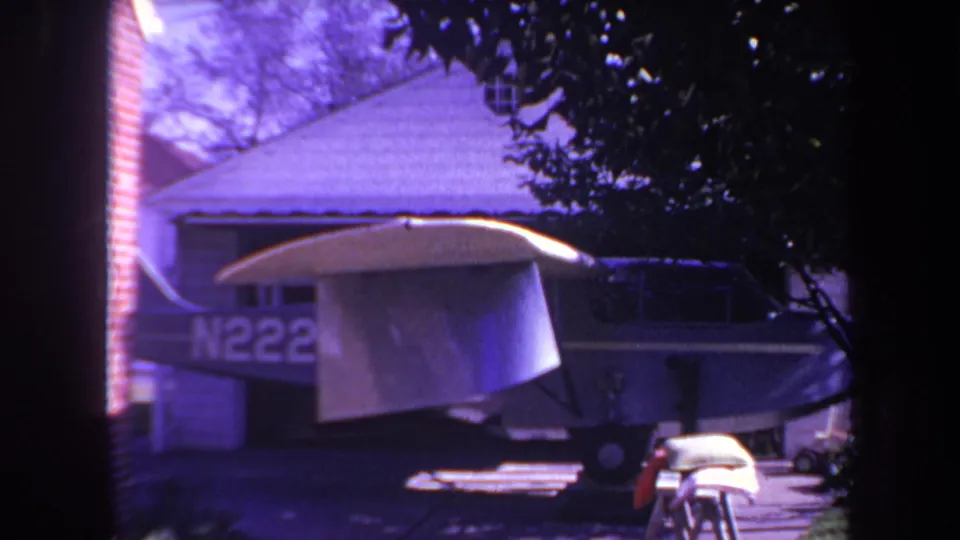 A small blue airplane with white wings sits in a residential driveway in the 1969 Yucaipa home-movie footage, with trees and a house structure behind it.