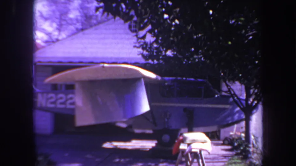 The 1969 8mm frame shows the airplane closer in view, with the white wing prominent, partial registration markings on the blue fuselage, and a driveway foreground.
