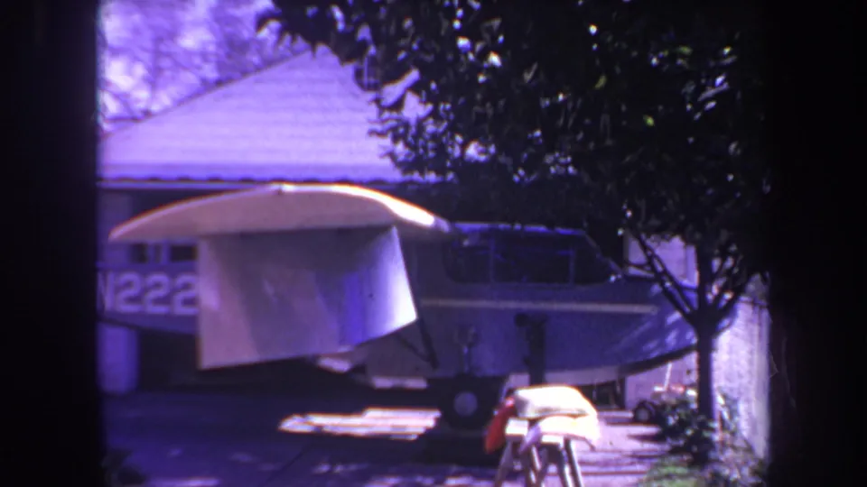Final 1969 Yucaipa home-movie frame showing the parked small airplane in partial shade, with white wings, blue fuselage, trees, and a residential structure visible.
