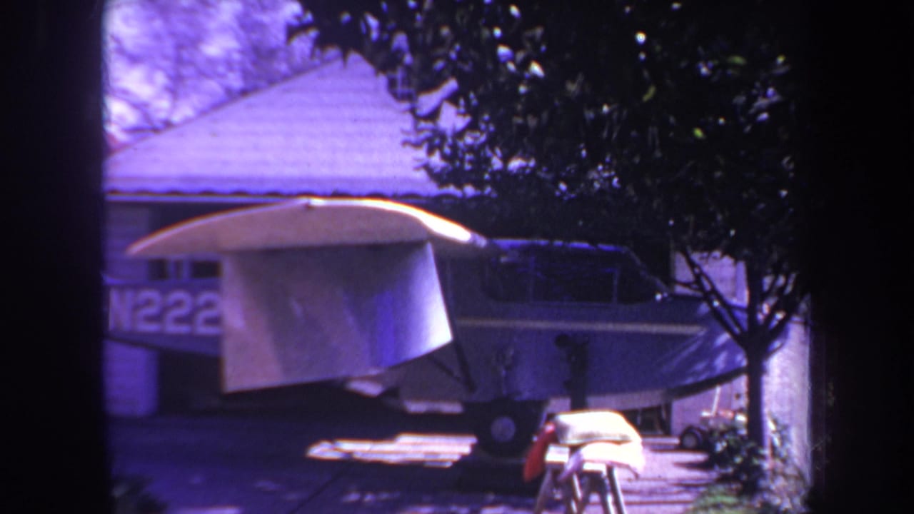 1969 8mm frame from Yucaipa, California, showing a small single-engine aircraft parked beside a house with a white tarp over its wing and trees framing the view.